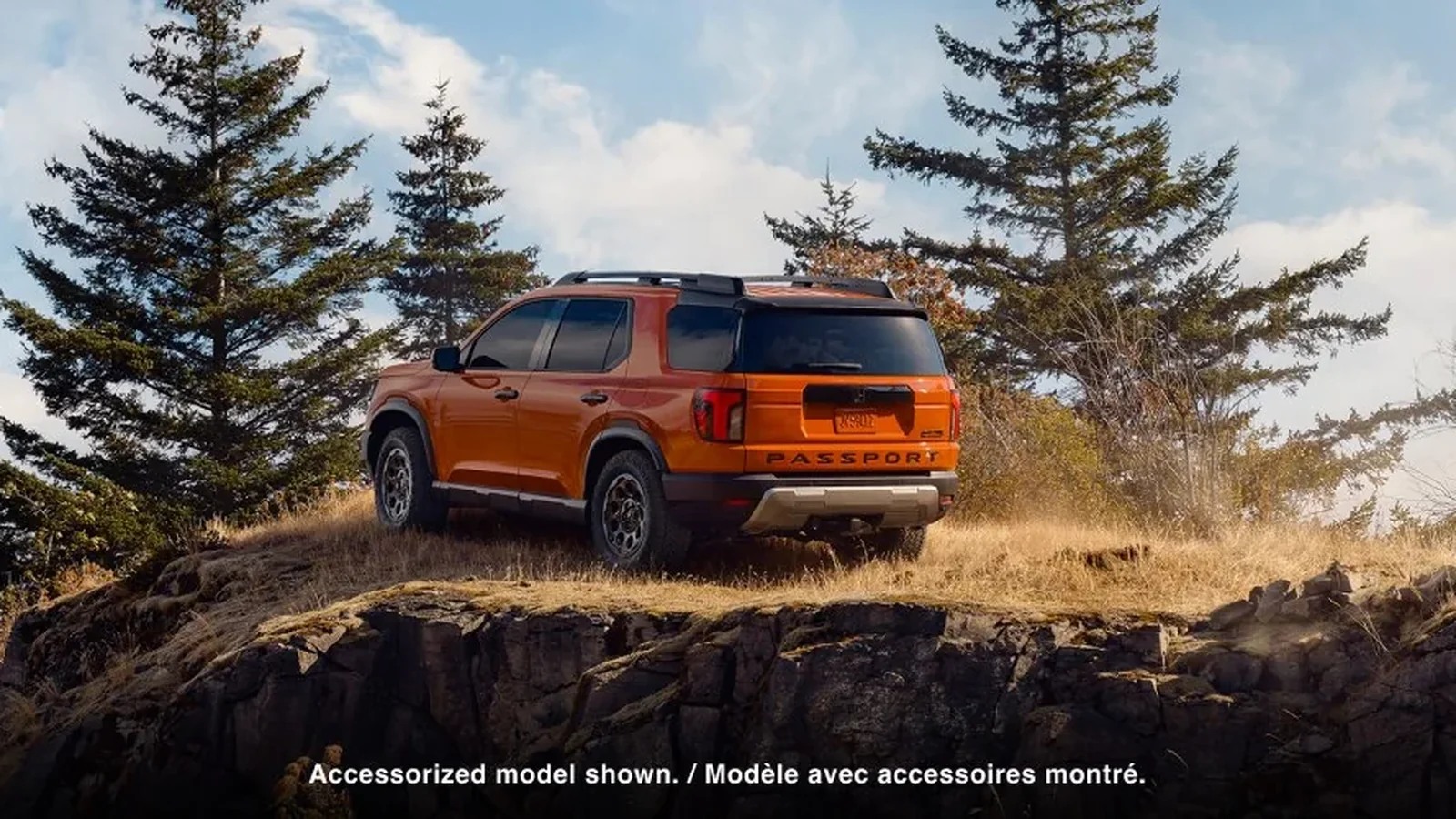 Orange Honda Passport parked on a rocky ledge among evergreens, seen from the rear three-quarter (2026 Canadian model, accessorized)